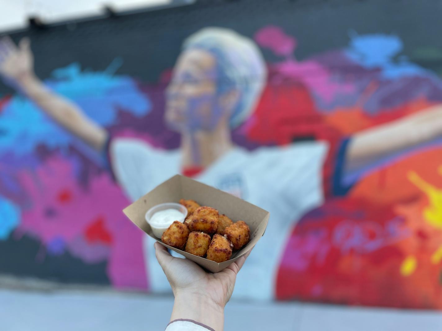 A basket of fried food is held up in front of a mural of soccer star Megan Rapinoe