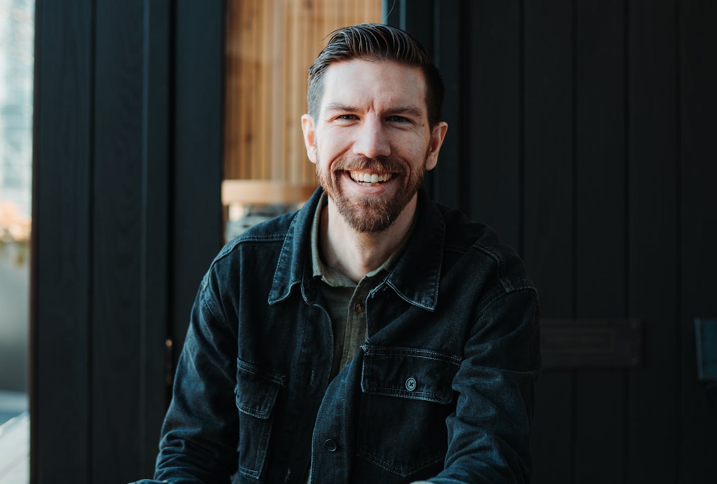 Cedar and Stone founder Justin Juntunen dressed in dark demin and collared shirt smiling in front of one of his company's saunas.