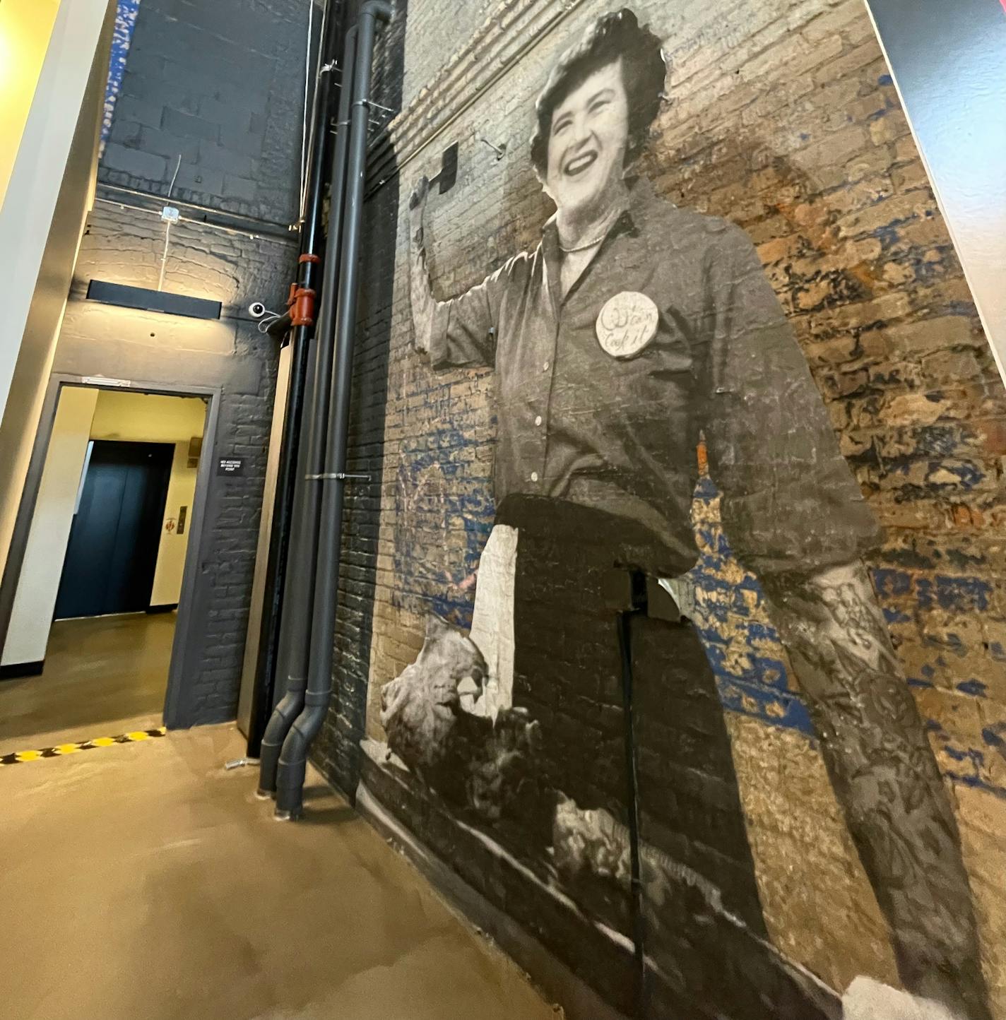 A black and white mural of Julia Child joyfully wielding a hammer, but her arms are decorated with full sleeves of tattoos.