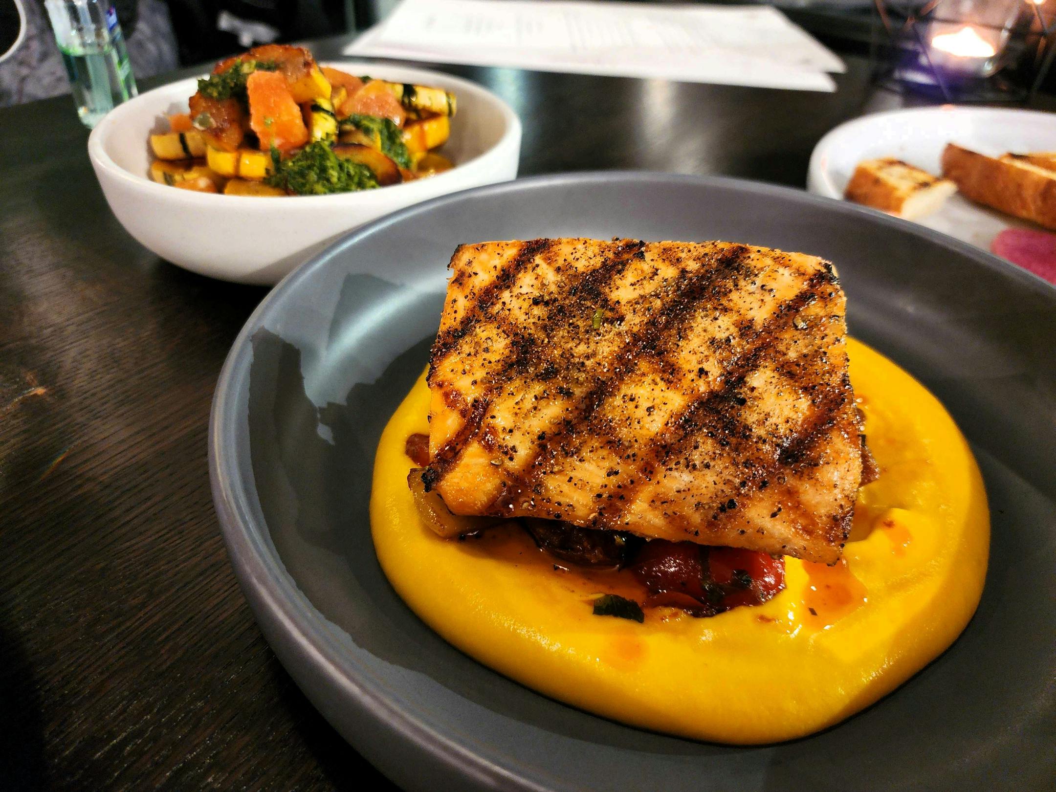A black plate with a square of salmon with diamond grill marks, on an orange-colored mousse
