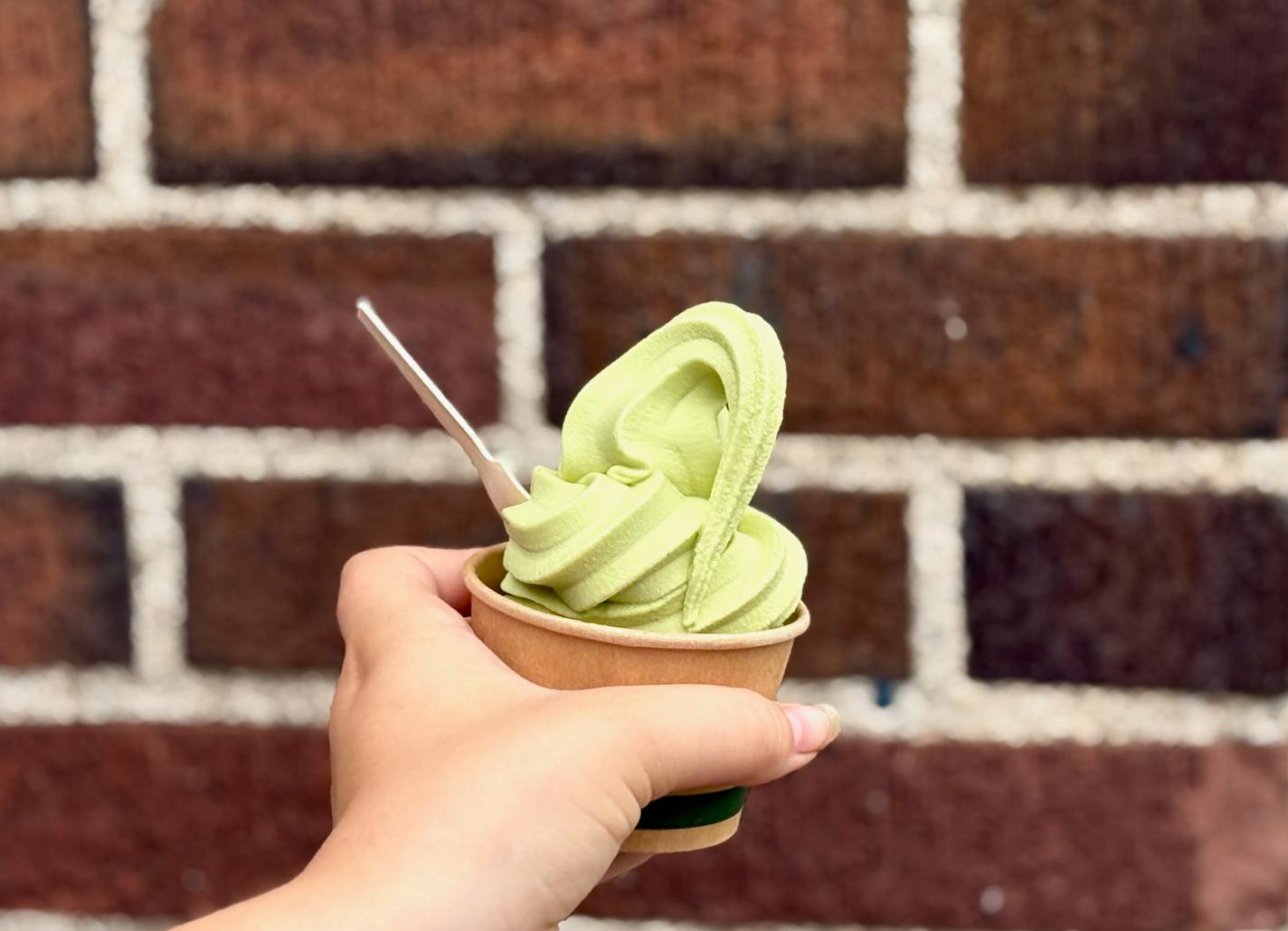 A cup of matcha soft serve held in front of the red brick outside the coffee shop.