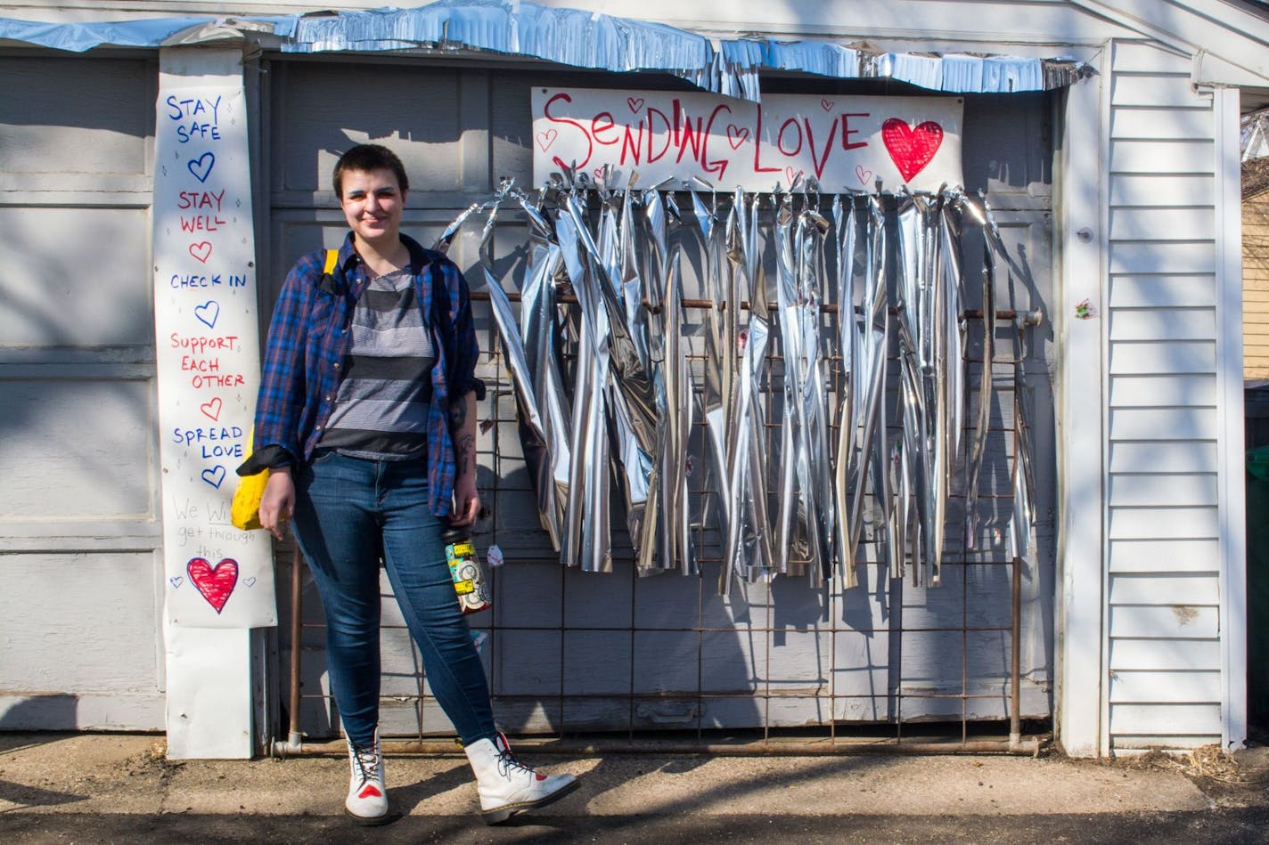 Art student Cedar Thomas smiles in front a collection of upbeat signs and decorations meant to cheeer neighborhood. Someone tore it all down and left a note accusing Thomas of not taking the pandemic seriously. Photo credit: Sophie Warrick