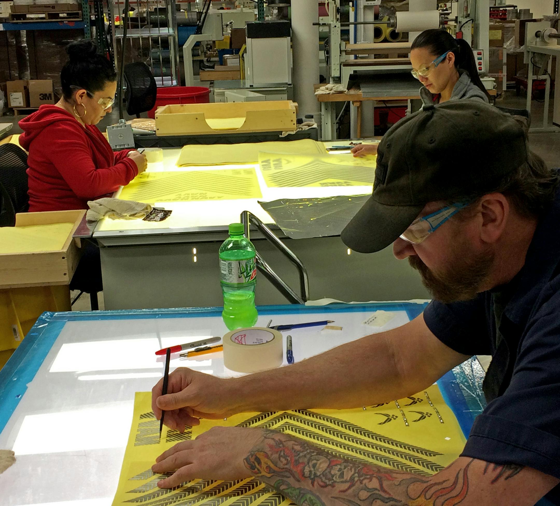 Photo 1: Mike Norve (man), Patricia Huerta (red) and Yeng Vang (gray) work on ‚Äúweeding, ‚Äù or removing unwanted pieces of reflective material with dental picks on light tables, on the ‚Äútransfer graphic‚Äù at Safe Reflections. The graphics will be laminated on Air Force or Navy uniforms.