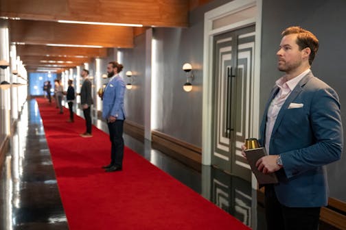 In a long hallway, men in suits stand along a red rug, waiting to be admitted into their private suites.