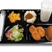 A black school lunch tray including tossed salad, chicken nuggets, roll, peaches, mashed potatoes and gravy with milk on a white background