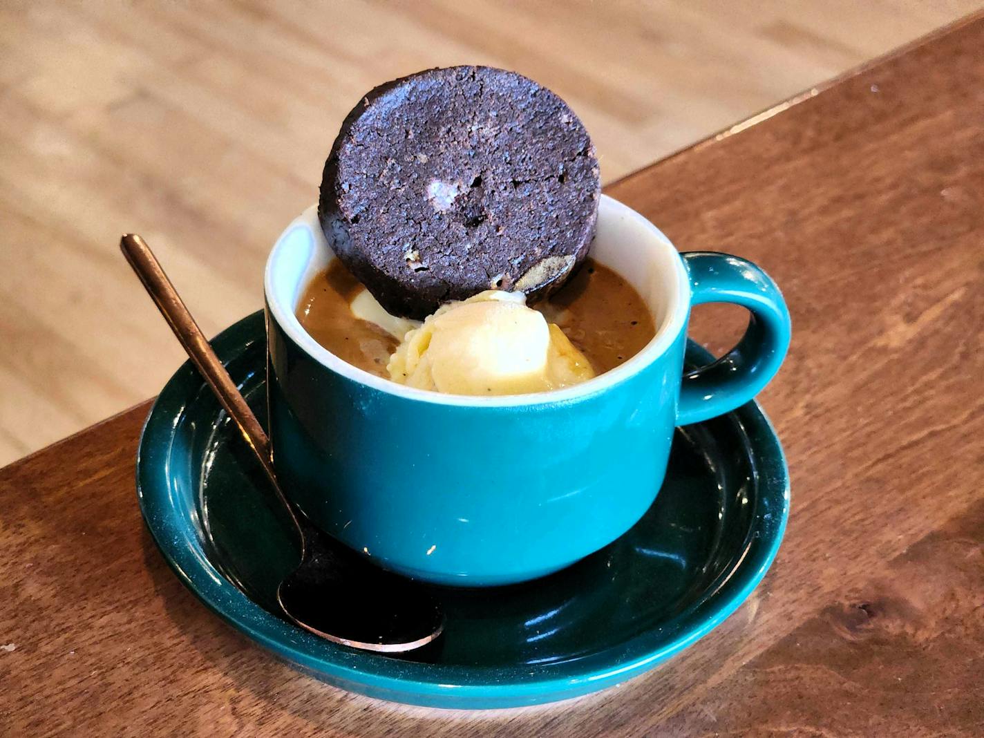 A wooden table holds a turquoise espresso mug and saucer. Inside is a pool of dark brown espresso with a small white scoop sticking out of it. A circular black cookie is resting on top. A gold spoon rests on the saucer.