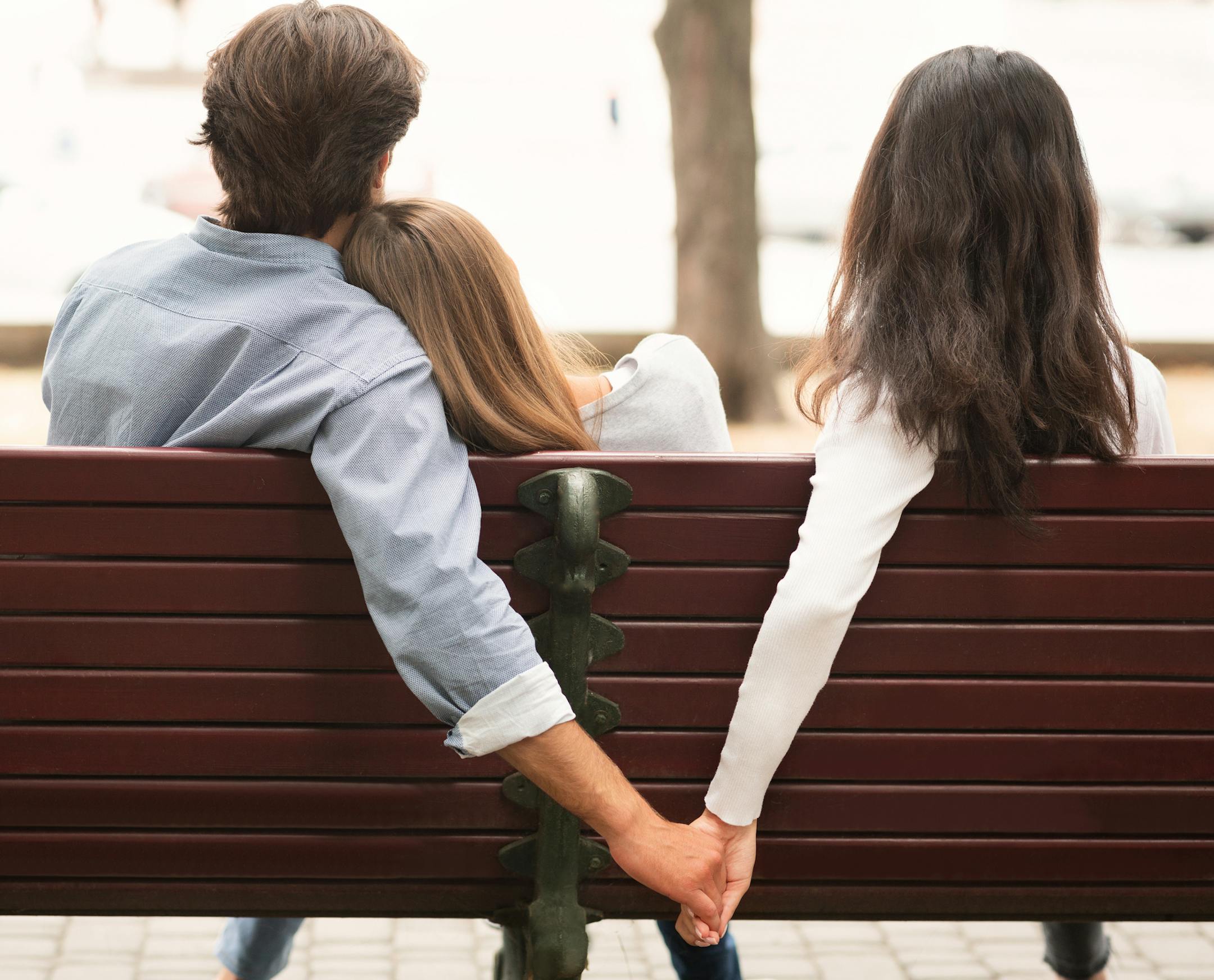 Love Triangle. Cheating Boyfriend Hugging Girlfriend Holding Hands With Her Girl Friend Sitting On Bench Together In Park Outdoor. Back-View