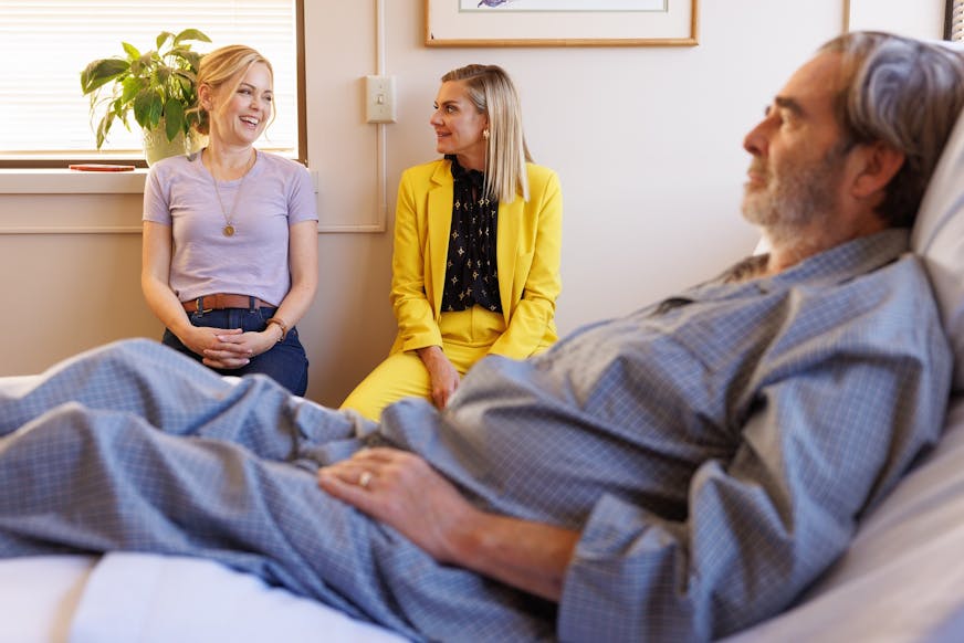 photo of an older man lying in a hospital bed in his pajamas, with two younger women seated next to the bed