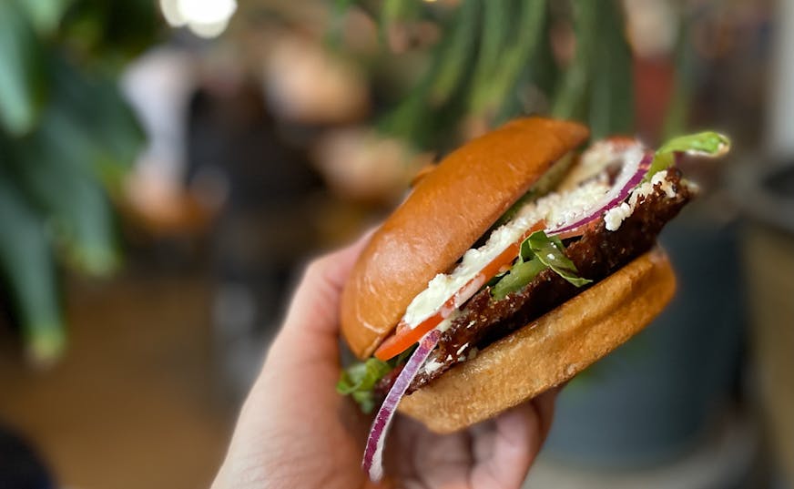 A thin burger patty with lots of lettuce, red onion, red tomato and crumbles of feta cheese on a bun.