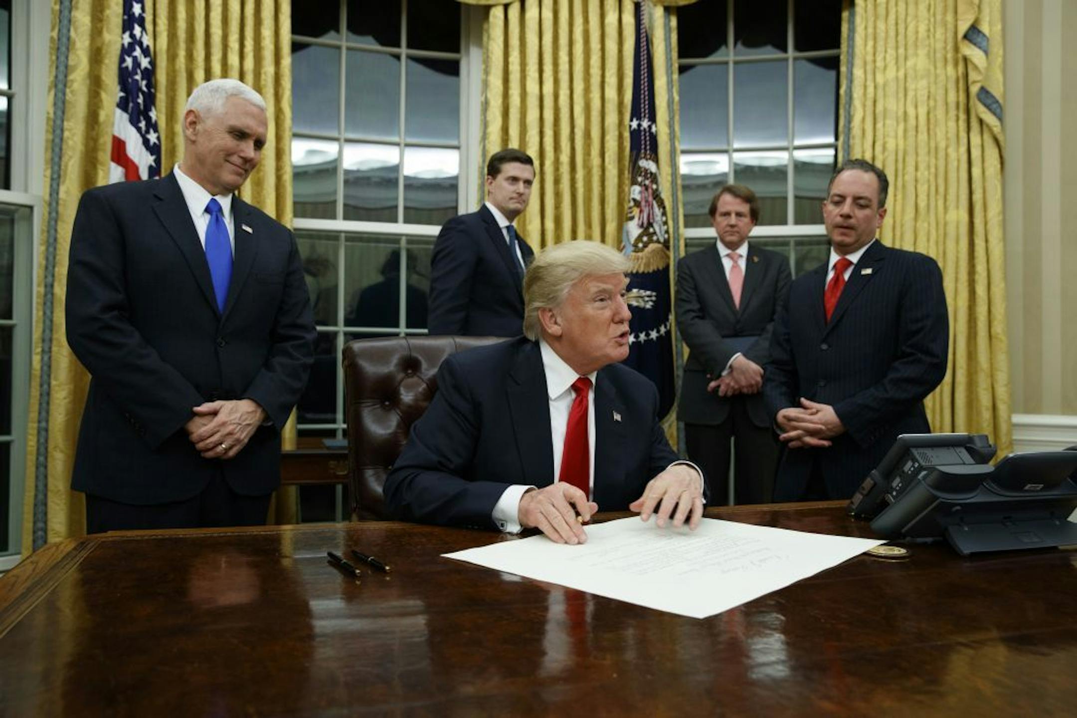 Vice President Mike Pence watches at left as President Donald Trump prepares to sign his first executive order, Friday, Jan. 20, 2017, in the Oval Office of the White House in Washington.