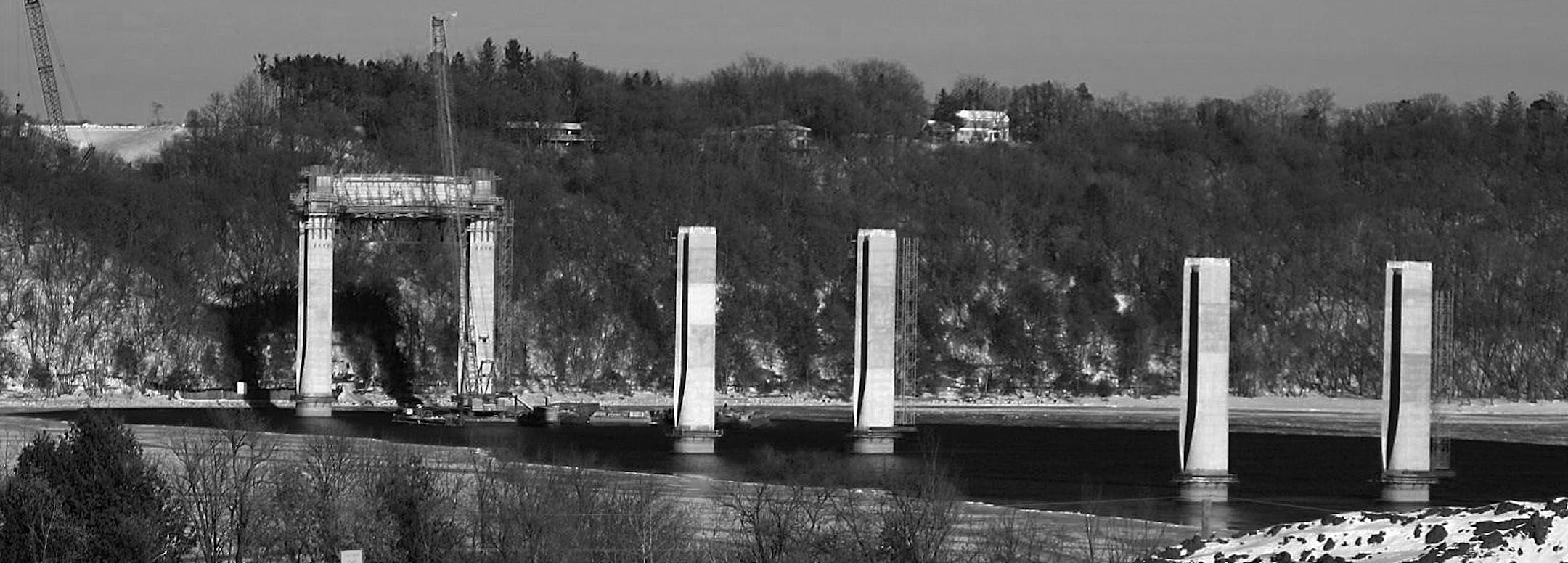 Sets of piers march east across the St. Croix River to the Wisconsin bluff. The piers stand about half as tall as the finished bridge with its cable supports.