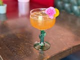 A margarita in a glass with a cactus shaped stem is a vibrant, orange-tinted color filled with ice and garnished with a pink mini carnation and round yellow flower.