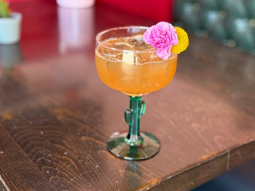 A margarita in a glass with a cactus shaped stem is a vibrant, orange-tinted color filled with ice and garnished with a pink mini carnation and round yellow flower.
