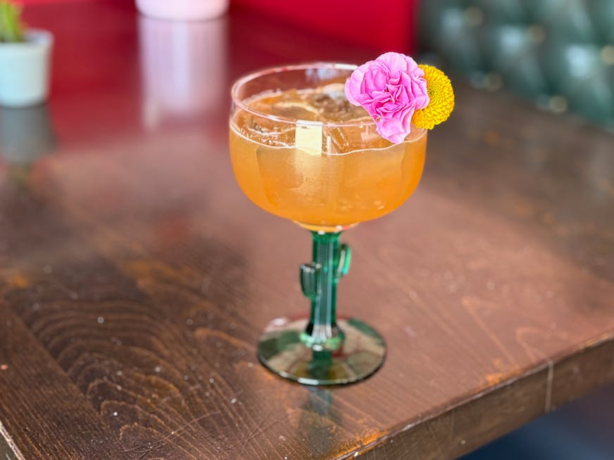 A margarita in a glass with a cactus shaped stem is a vibrant, orange-tinted color filled with ice and garnished with a pink mini carnation and round yellow flower.