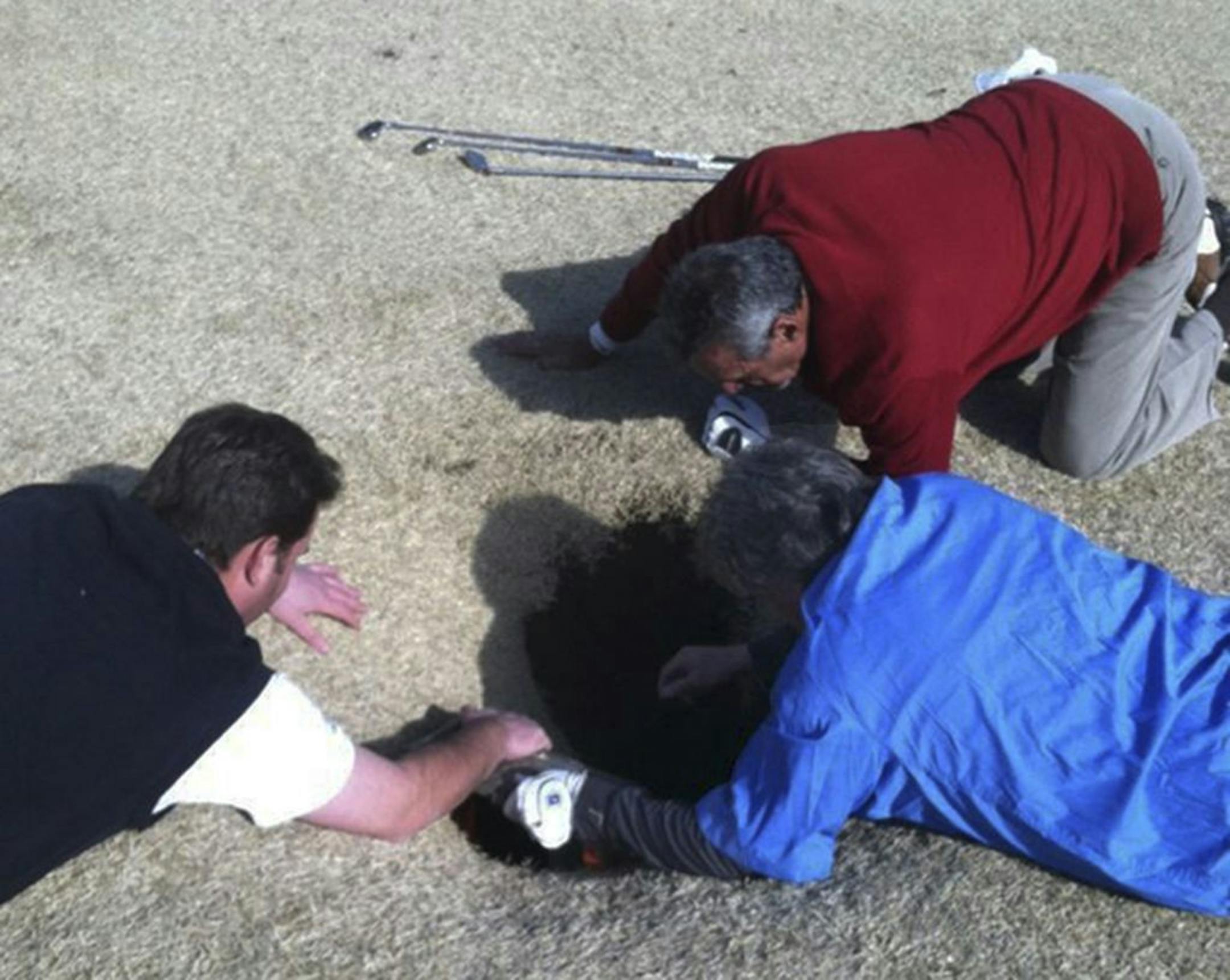 In this cell phone image taken March 8, 2013 and provided by golfmanna.com, Hank Martinez, top, Ed Magaletta, right, and Russ Nobbe, look into an 18-foot-deep and 10-foot- wide sinkhole that golfer Mark Minhal fell into while playing golf at the Annbriar Golf Course in Waterloo, Ill. Mihal, 43, a mortgage broker from Creve Coeur, Mo., was hoisted to safety with a rope and suffered a dislocated shoulder.