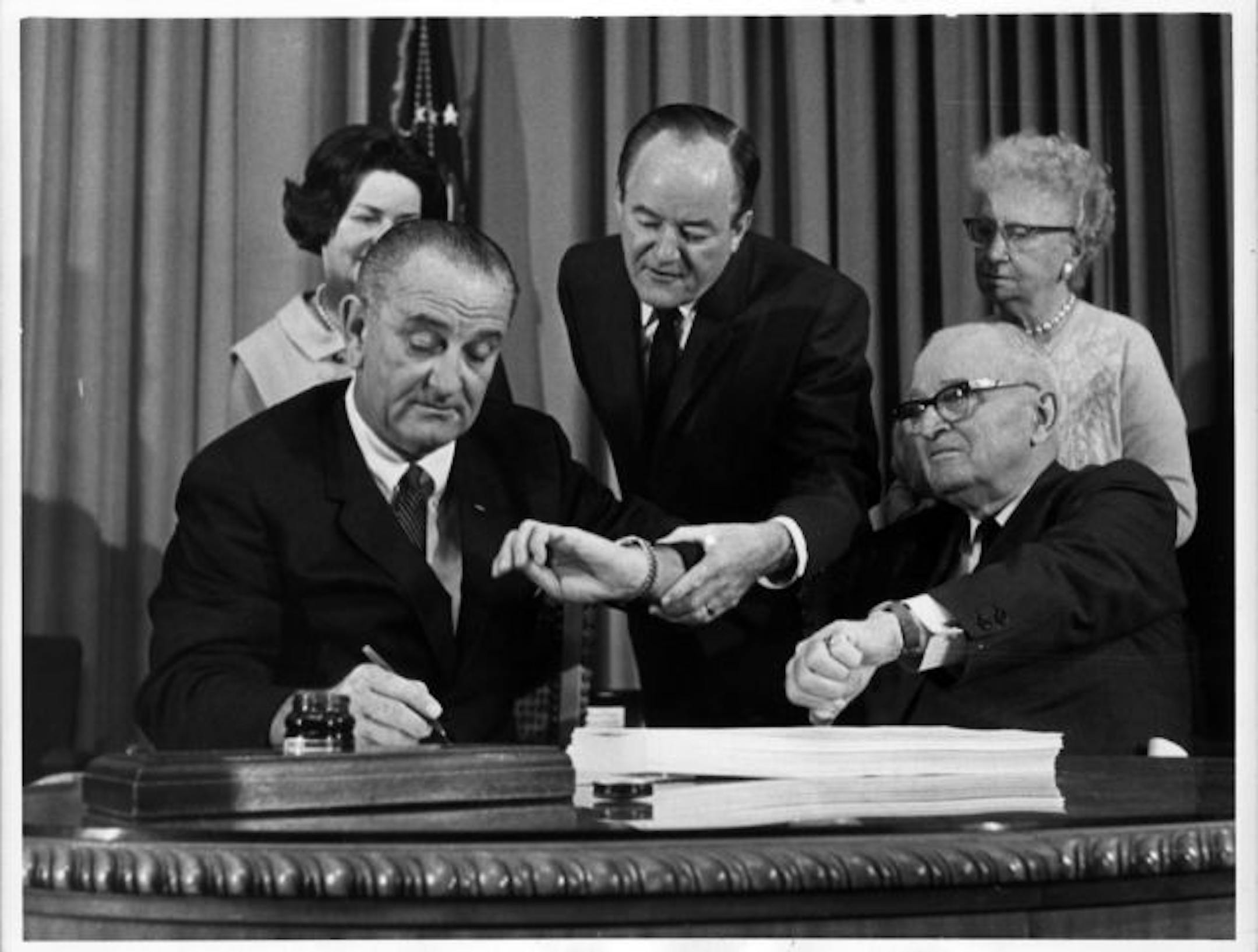President Lyndon Johnson signed the Medicare bill July 30, 1965, as Vice President Hubert Humphrey and former President Harry Truman checked the time of the signing in ceremonies at the Truman Library in Independence, Mo. Lady Bird Johnson and Bess Truman stood behind their husbands. / File photo.