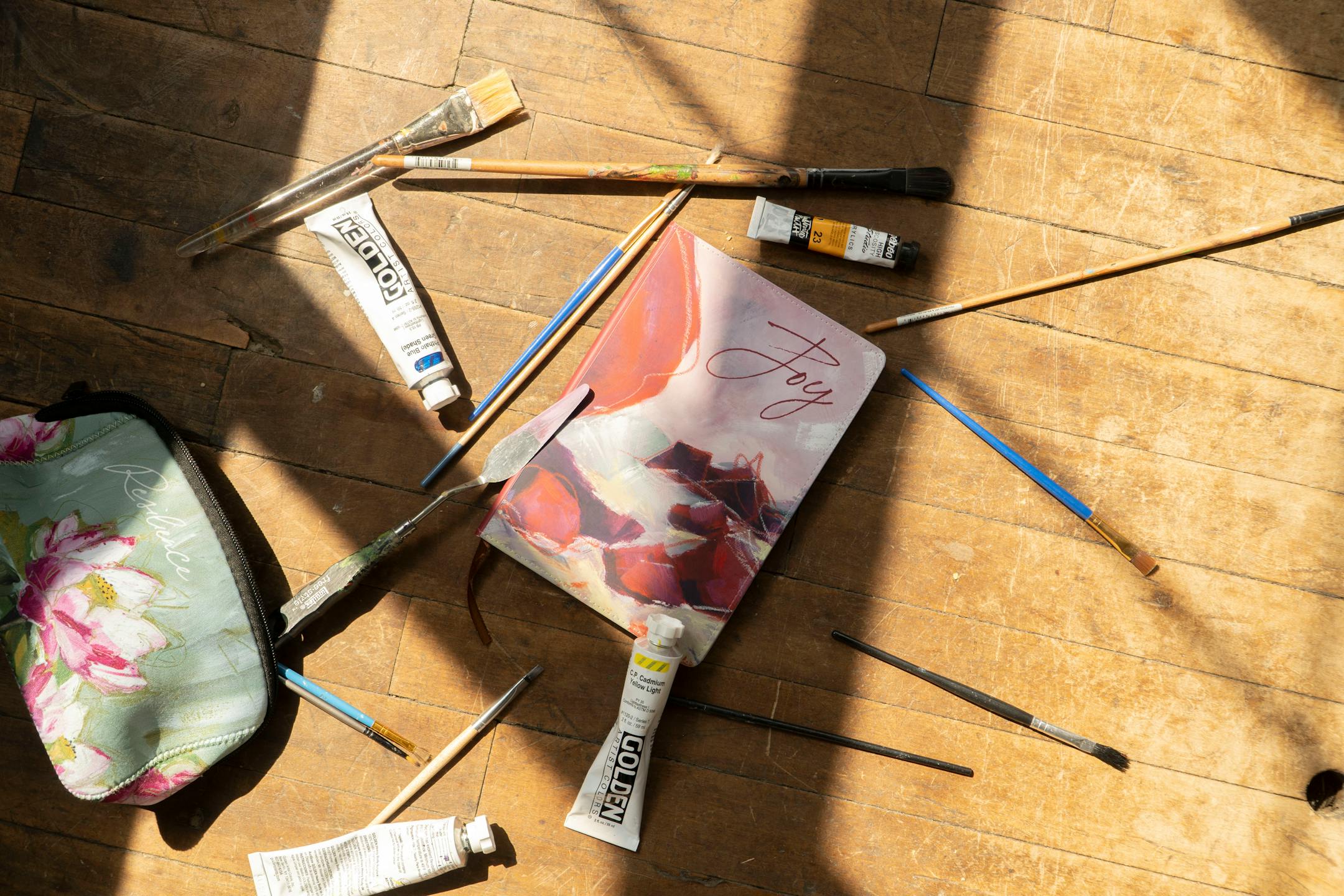 Scattered across the wooden floor amid a handful of paintbrushes are a makeup bag bearing the image of a lotus flower and a writing journal depicting a rocky shore.