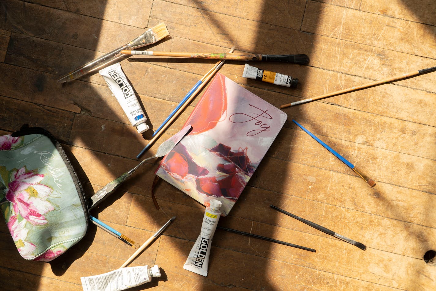 Scattered across the wooden floor amid a handful of paintbrushes are a makeup bag bearing the image of a lotus flower and a writing journal depicting a rocky shore.