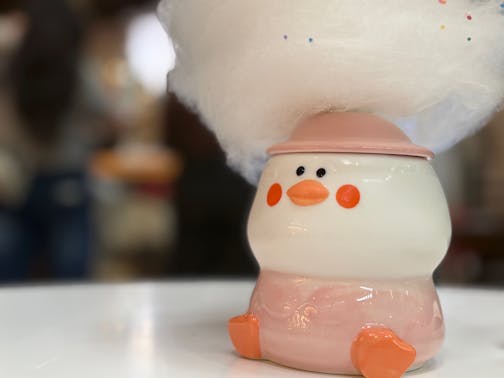 A squat ceramic duck mug with rosy cheeks and little feetsies has a cone hat and a swirl of sprinkle-dotted cotton candy coming out of his head. Sitting on a white table top.