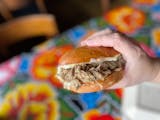 A hand holds a golden bun filled with juicy-looking meat, topped with horseradish cream that's starting to drop, over a bright blue, floral oilcloth covered table.