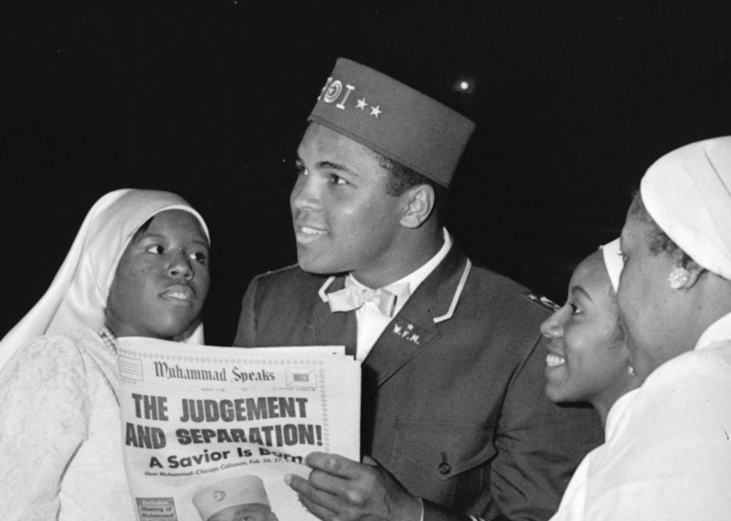 Muhammad Ali, world heavyweight champion, wears a uniform of the Black Muslim organization as he attends their convention, February 26, 1966, in Chicago. He is talking with Sisters of Islam, female members of the group who wear the habit of their order. The newspaper held by Ali carries photo of Elijah Muhammad, head of the Black Muslims. (AP Photo) ORG XMIT: C12