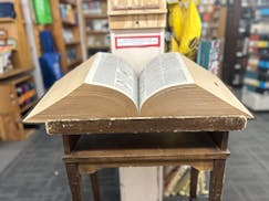 A corduroy bound dictionary weighing over 25 pounds and measuring 9 inches tall sits on a rickety wooden end table in a bookstore.