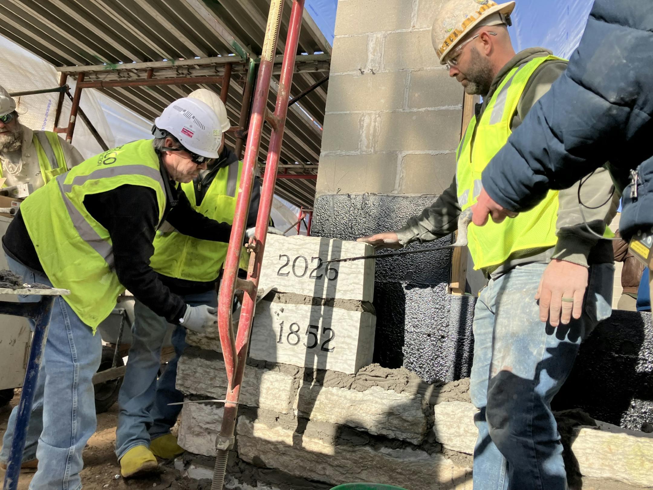 Three construction workers in white hard hats, neon yellow vests and blue jeans stack two limestone blocks and apply mortar. The blocks are engraved with the years 1852 and 2026.