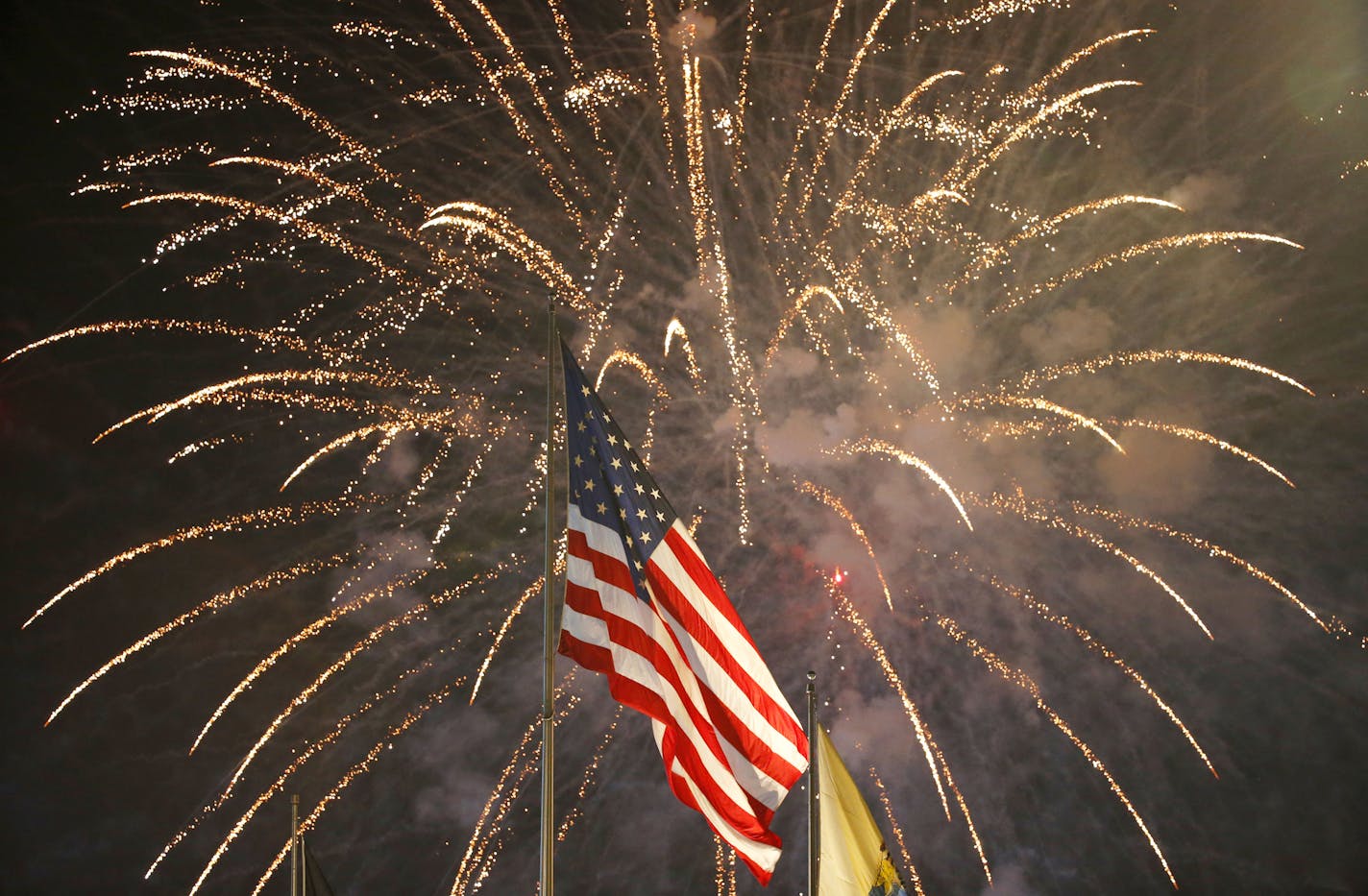 FILE - In this July 4, 2015, file photo, fireworks explode behind a United States flag during a Fourth of July celebration at State Fair Meadowlands in East Rutherford, N.J. With fewer professional celebrations on July 4, 2020, many Americans are bound to shoot off fireworks in backyards and at block parties. And they already are: Sales have been booming. (AP Photo/Julio Cortez, File)