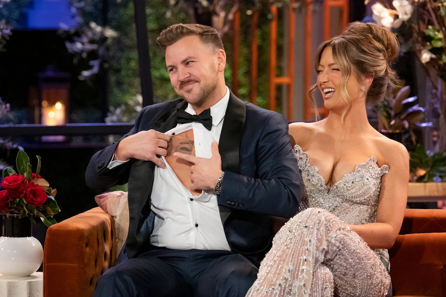 A man in a tuxedo opens his shirt and points to a tattoo of a woman's eyes on his chest while his wife in a sparkly dress laughs beside him.