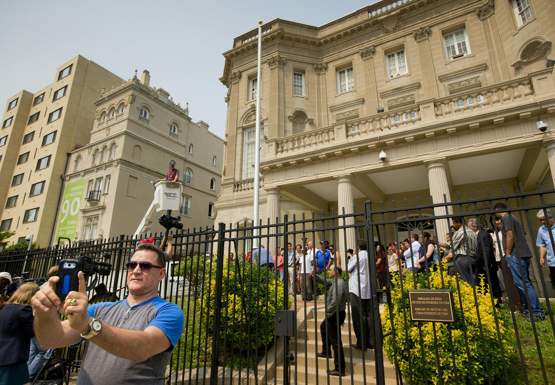 Fernando Rodriguez, left, uses his cellphone to take a selfie as workers from Eastern Shores Flagpoles raise a flagpole at the Cuban Interest Section in Washington in preparation for re-opening of embassies in Havana and Washington, Wednesday, June 10, 2015. Rodriguez from Lancaster, Pa., but originally from Mantanzas, Cuba, was visiting Washington to get his passport renewed and wasn't aware of the event until he saw all the commotion. Currently the Cuban Interest Section in US is under the aus