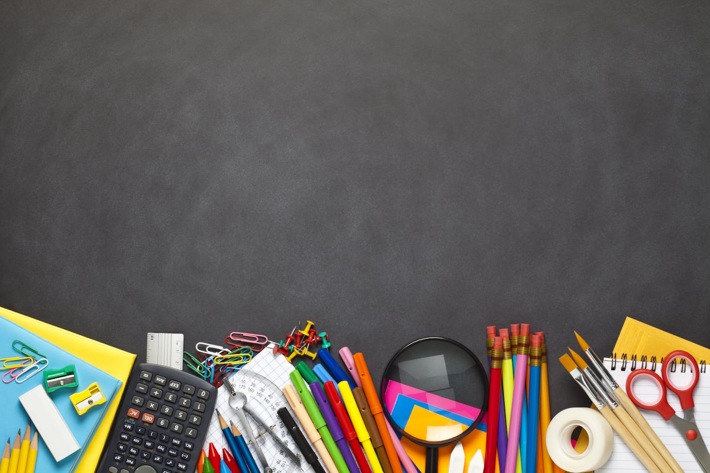 Black chalkboard with school supplies coming from the bottom border of the frame. The composition includes paper clips, yellow pencils, note pad, loupe, adhesive tape, paint brushes, crayons, pencil sharpener, scissors, compass, ruler, calculator and pushpins. Useful copy space left at the center and top of the frame. High angle view DSRL studio photo taken with Canon EOS 5D Mk II and Canon EF 100mm f/2.8L Macro IS USM