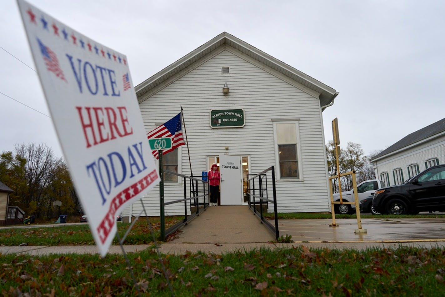 "Vote Here Today" sign in front of small white frame town hall with person in red jacket leaving building.