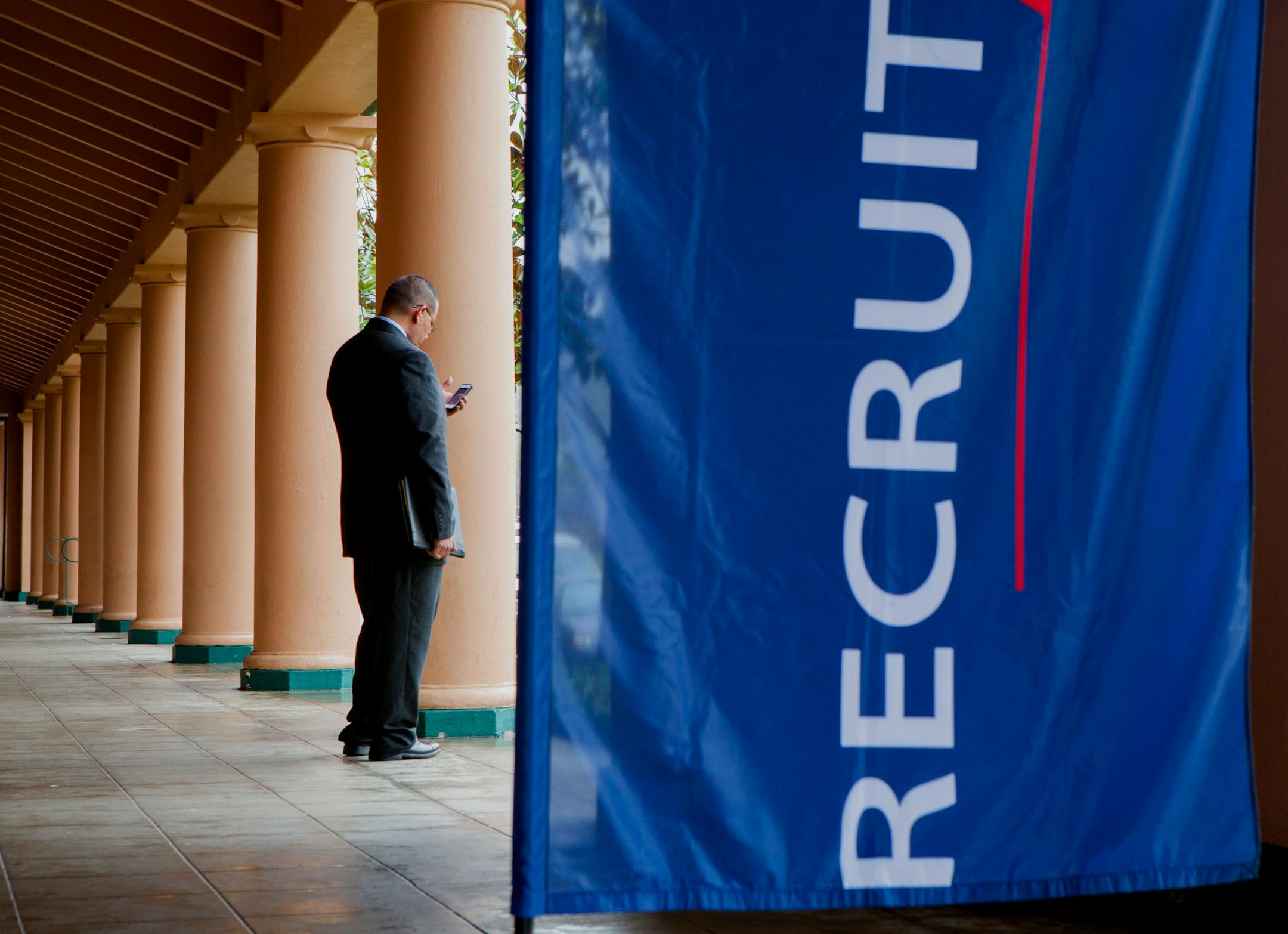 A job seeker checks his mobile phone outside the Recruit Military veteran job fair in San Diego, California, U.S., on Thursday, Feb. 27, 2014. More Americans than forecast filed applications for unemployment benefits last week, a sign the labor market is improving in fits and starts. Photographer: Sam Hodgson/Bloomberg