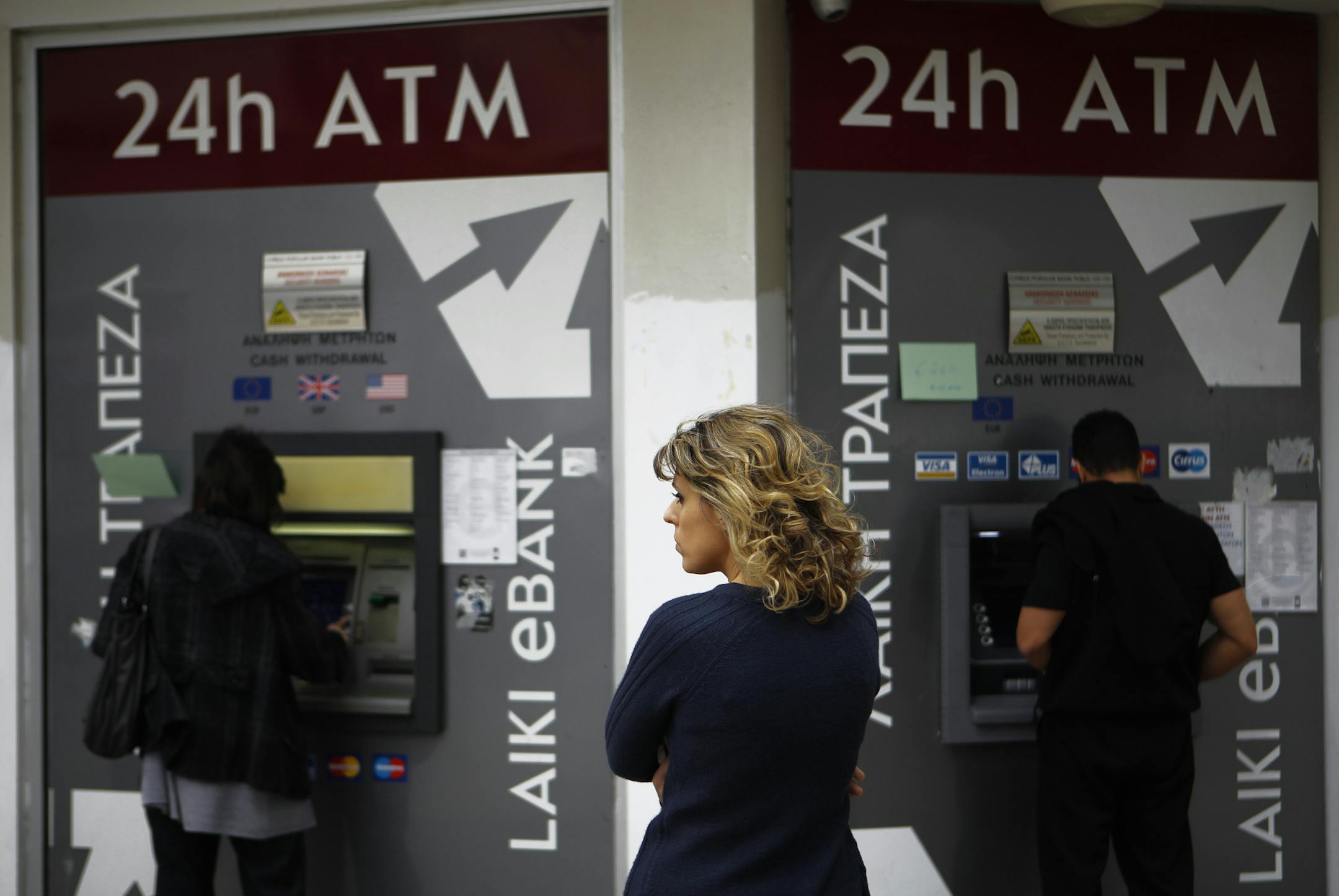 A woman waits as two people use the ATM machines in central capital Nicosia, Cyprus, Friday, March 22, 2013. Cypriot authorities were putting the final touches Friday to a plan they hope will convince international lenders to provide the money the country urgently needs to avoid bankruptcy within days. (AP Photo/Petros Karadjias)