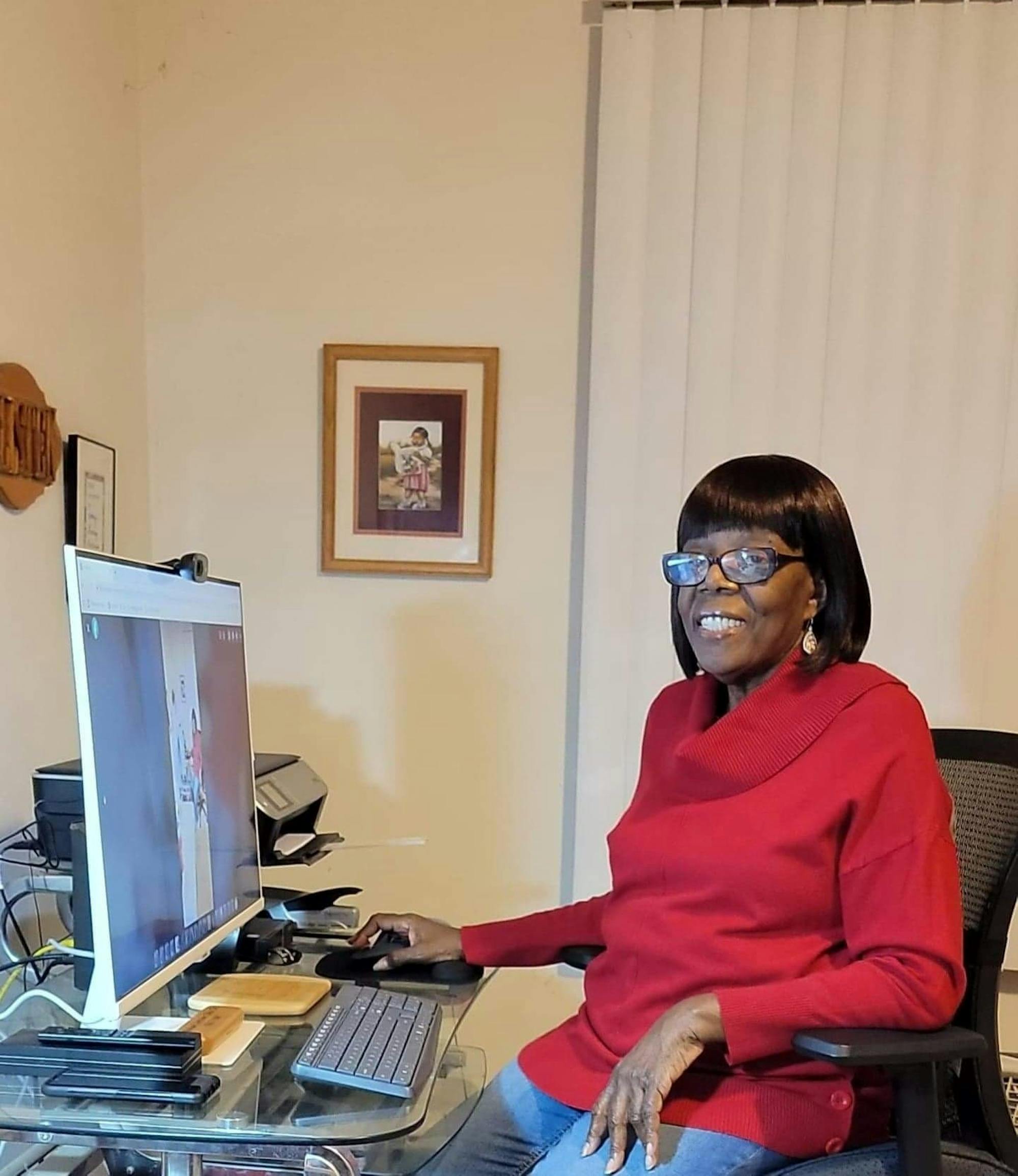 A woman at a desk with a computer on it smiles toward the camera.