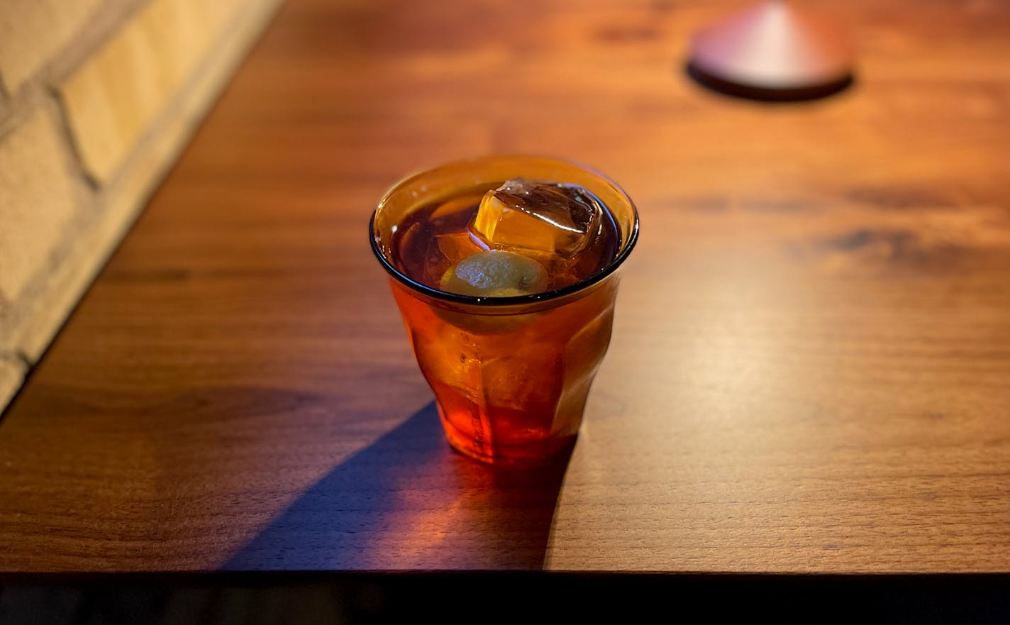 A lowball on a wooden table with a clear cocktail bobbing on top and a small orange peel garnish. There's a dim light that casts a purplish colored cast.