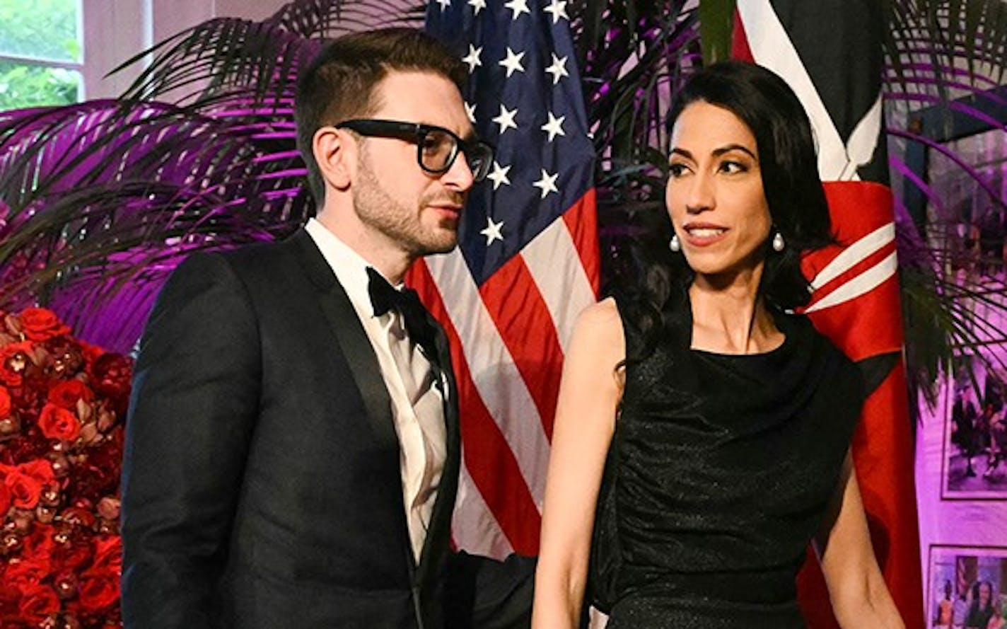 Alex Soros in tuxedo and Huma Abedin in black sleeveless gown with background of pink and red flowers in vases and a U.S. and other flags displayed.