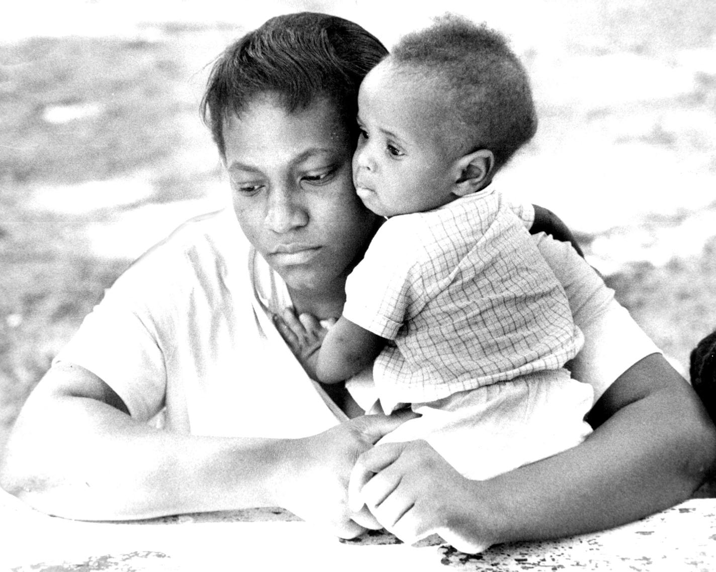 In a file photo from 1962, Betty Beal, a young Black woman from Louisiana, sits at a picnic table, cheek to cheek with her baby boy.