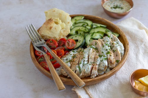 A plate of grilled chicken topped with herb yogurt sauce served with cherry tomatoes and cucumbers and fresh pita.