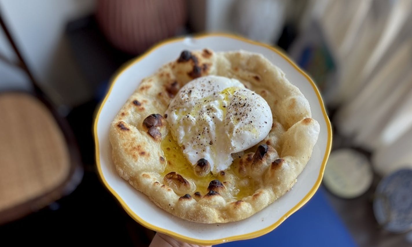 A white plate holds a round and bubbly flatbread with a white orb of cheese at its center