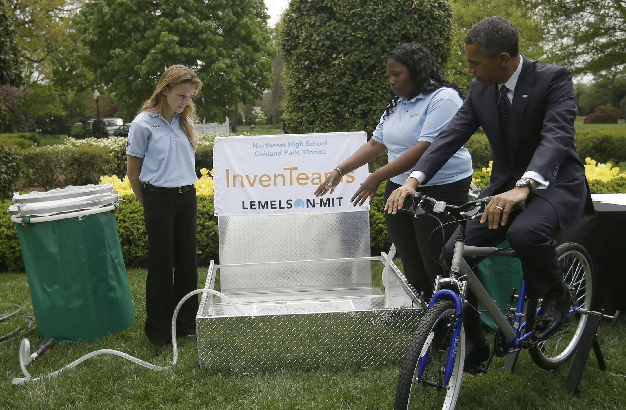 President Barack Obama pedals bicycle-powered emergency water-sanitation station for Payton Karr, 16, left, and Kiona Elliott, 18, center, both from Oakland Park, Fla., to help demonstrate their invention, Monday, April 22, 2103, in the East Garden of the White House in Washington, where the president hosted the White House Science Fair to celebrate the student winners of a broad range of science, technology, engineering and math (STEM) competitions from across the country. The bicycle filters E