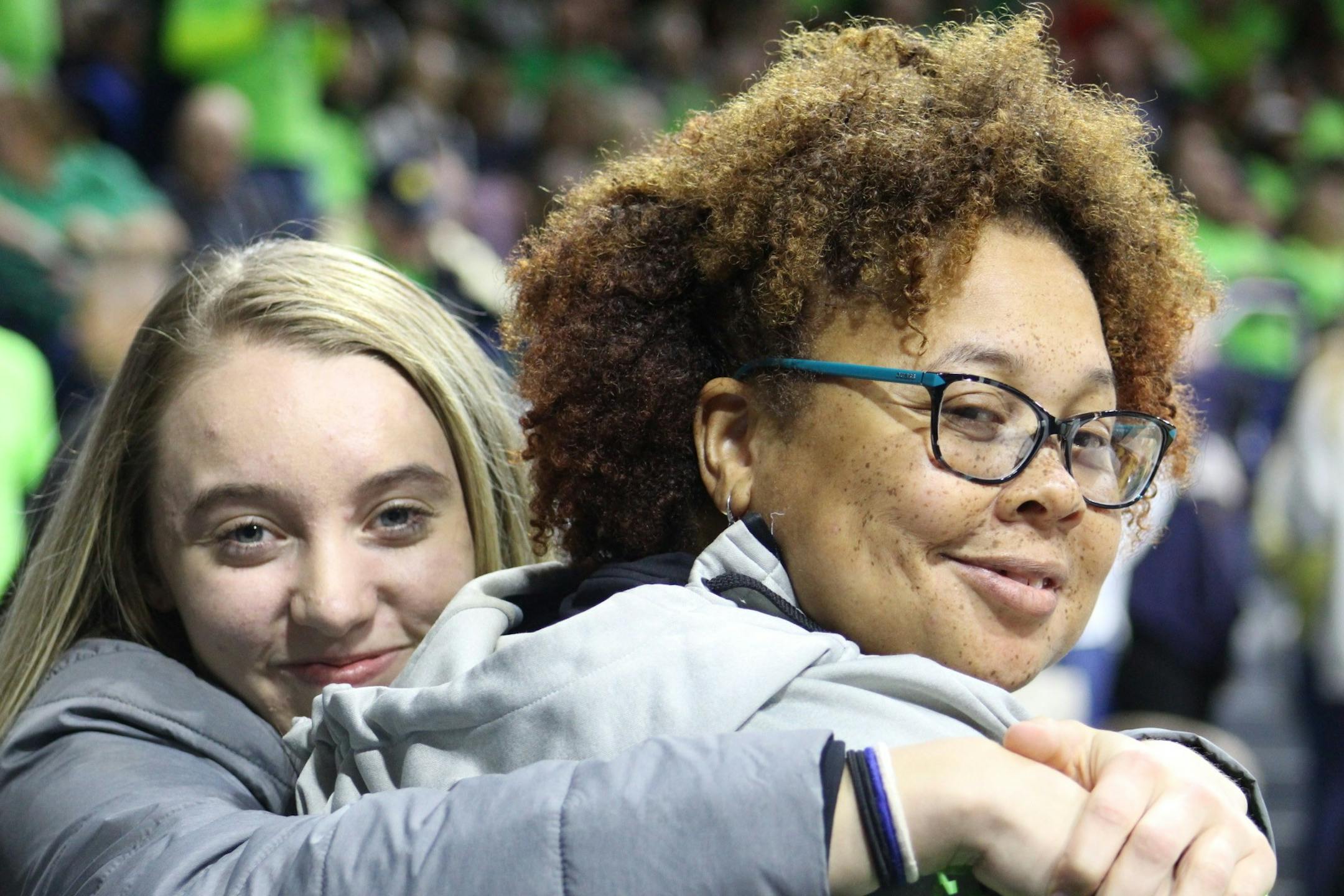 A young blond woman wraps her arms around an African American woman with glasses at a basketball game. Both are smiling and looking at the camera.