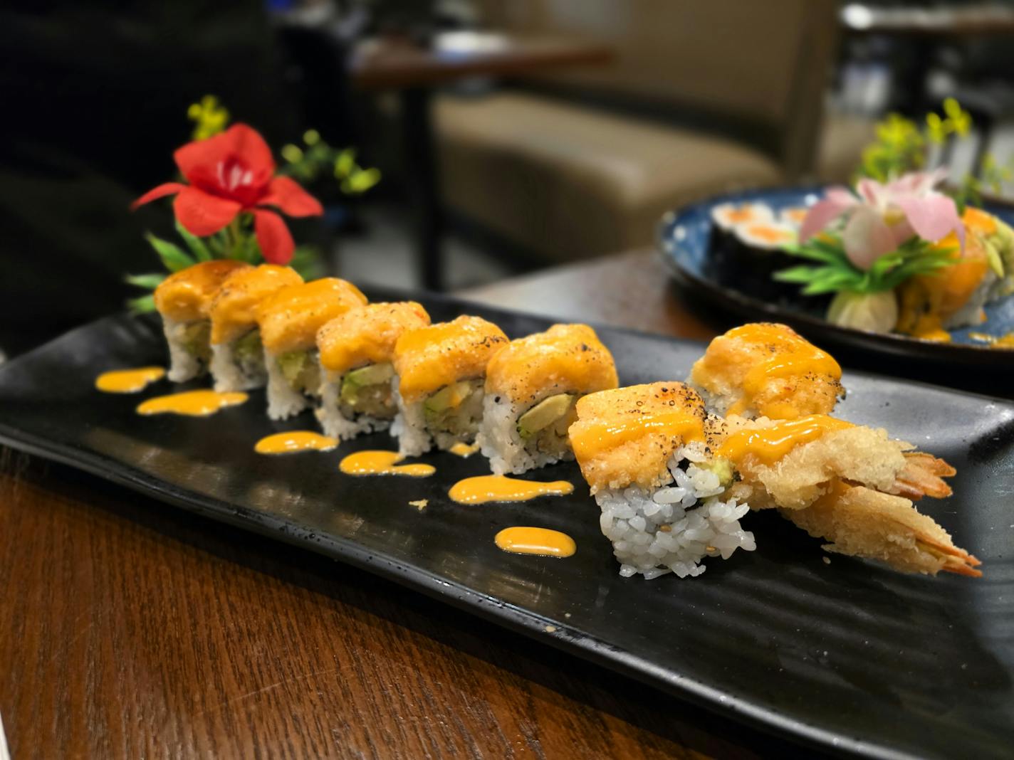 A sushi roll with orange-colored sauce drizzled on top, on a table in front of a flower