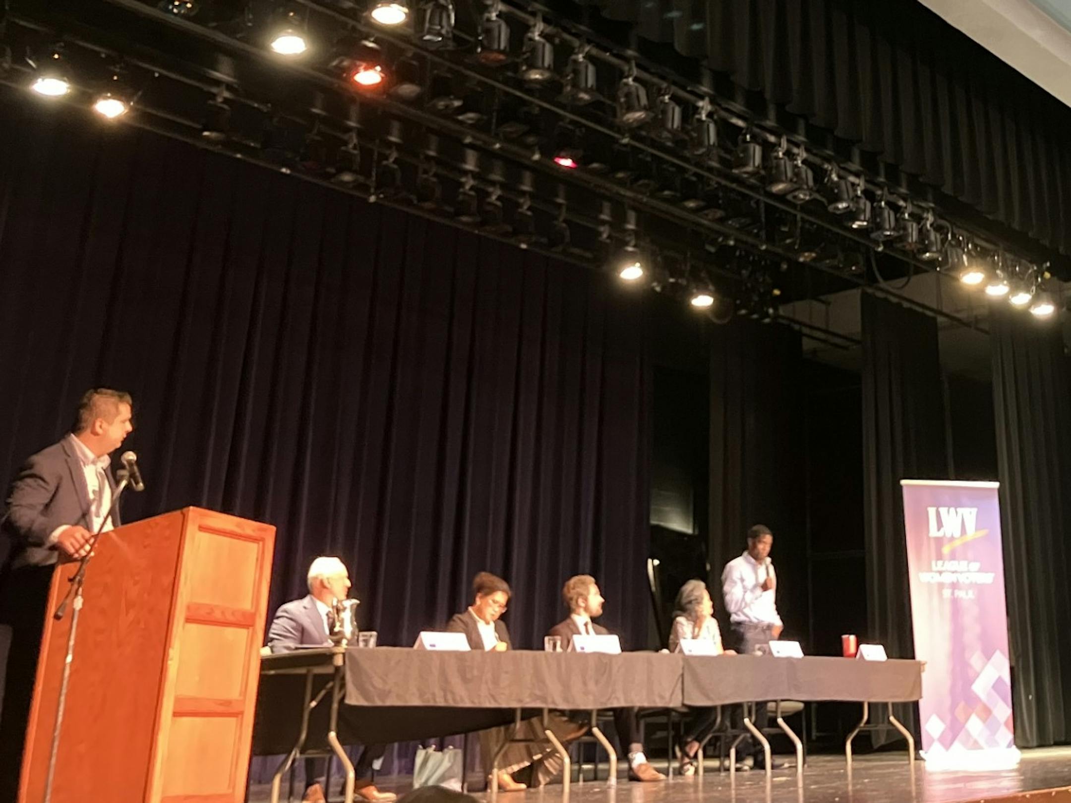 Mike Hilborn, State Rep. Kaohly Vang Her, Adam Dullinger and Yan Chen sit at a long table on a stage while Melvin Carter stands at the end of the table and speaks into a microphone. A moderator stands at a podium in the foreground.