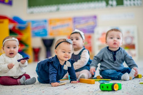 A 1 year old girl crawls on the carpet while smiling. A group of babies sitting next to her look around curiously. They are in a colorful daycare environment.
