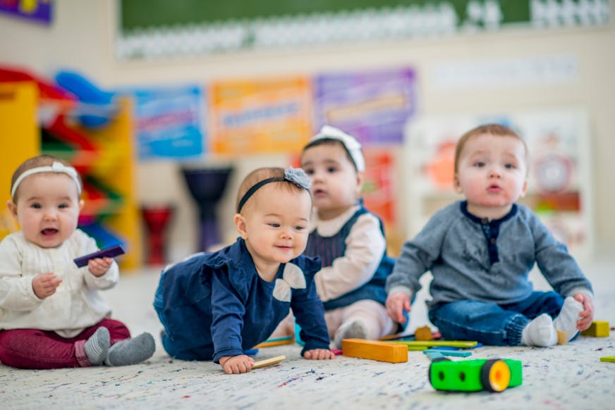 A 1 year old girl crawls on the carpet while smiling. A group of babies sitting next to her look around curiously. They are in a colorful daycare environment.