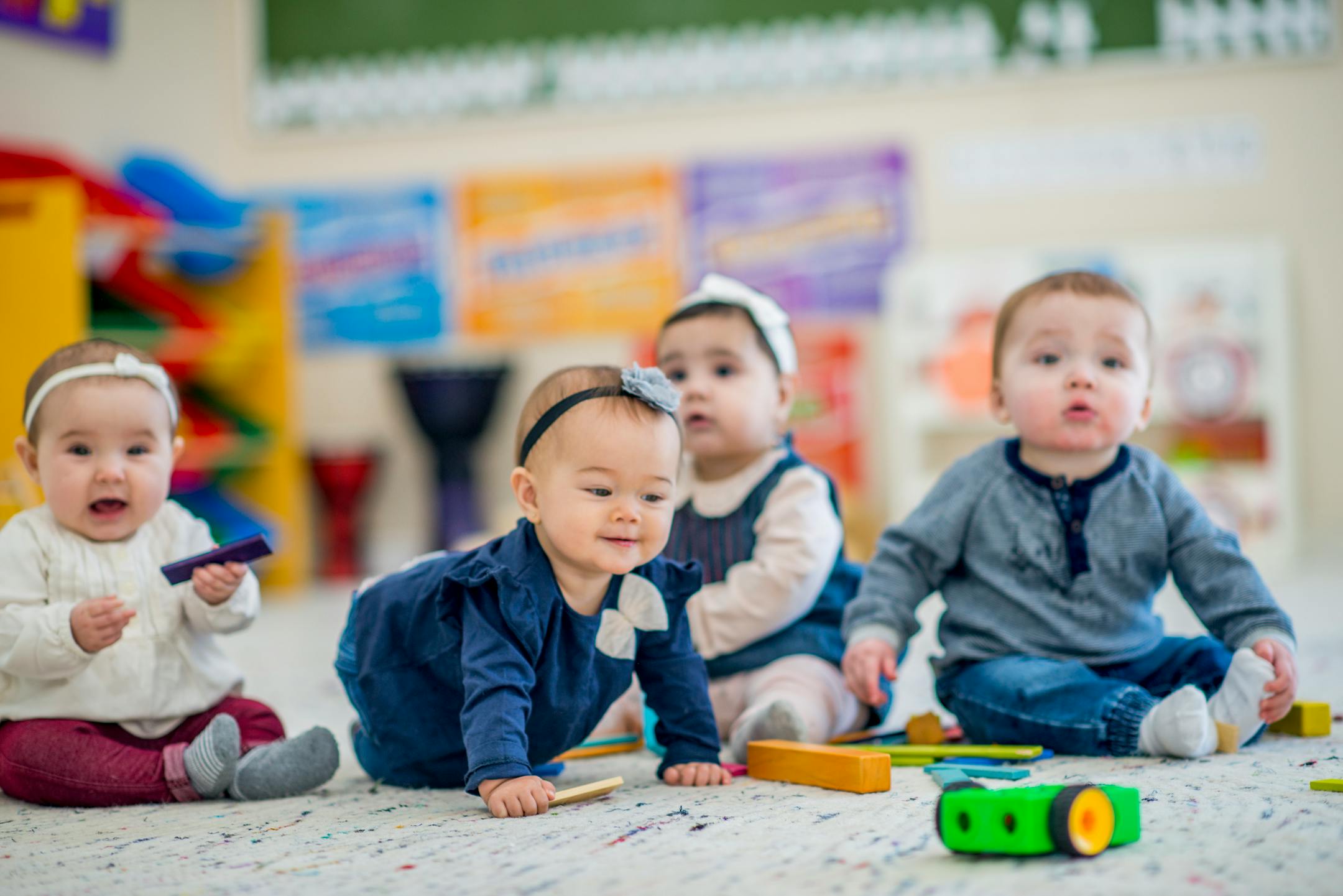 A 1 year old girl crawls on the carpet while smiling. A group of babies sitting next to her look around curiously. They are in a colorful daycare environment.