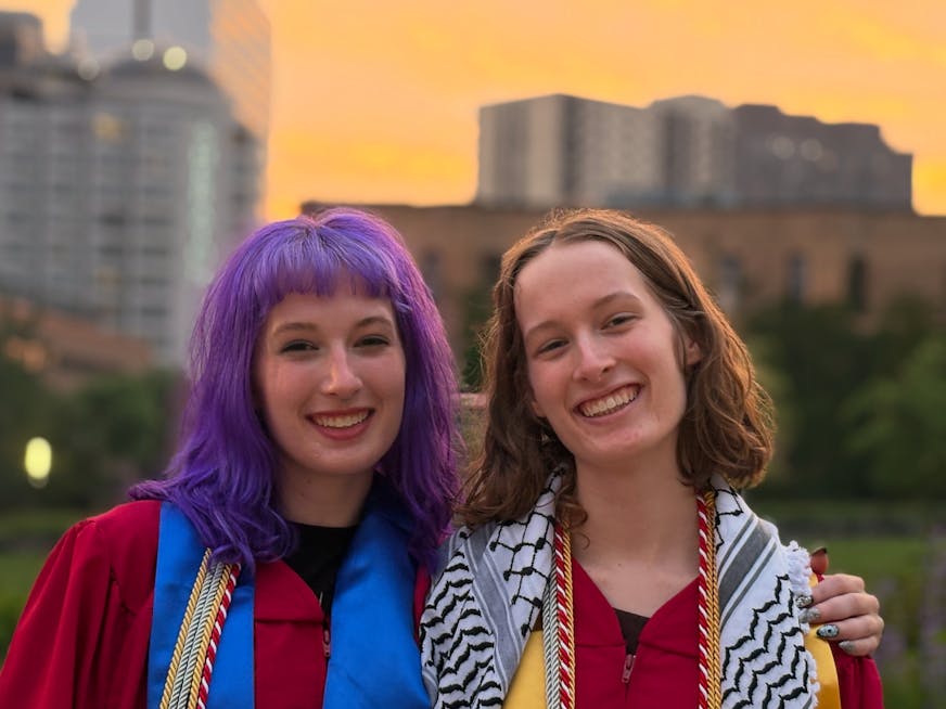 Identical twins - one with purple hair and colorful scarf, the other with a black and white patterned scarf - smile for the camera in front of tall buildings and a hazy sunset sky.