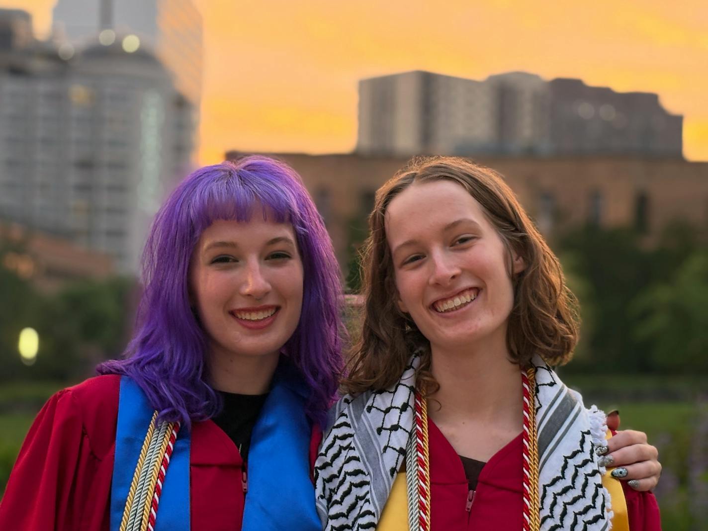 Identical twins - one with purple hair and colorful scarf, the other with a black and white patterned scarf - smile for the camera in front of tall buildings and a hazy sunset sky.