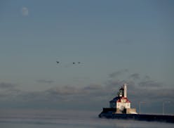 The moon rises in the sky as ducks fly by a light house in Duluth harbor in December 2017.